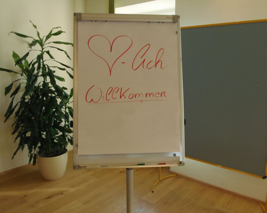 Ein Flipchart in einem Raum mit der Aufschrift „V-Ich Willkommen“ in roter Schrift und einem großen Herz darüber. Daneben steht eine große Topfpflanze.