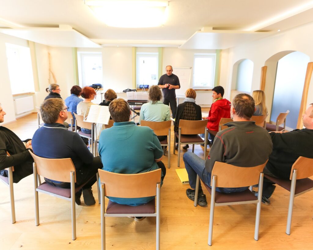 Ein Seminarleiter steht vor einer Gruppe von Erwachsenen, die auf Stühlen in einem hellen Raum sitzen und ihm aufmerksam zuhören.