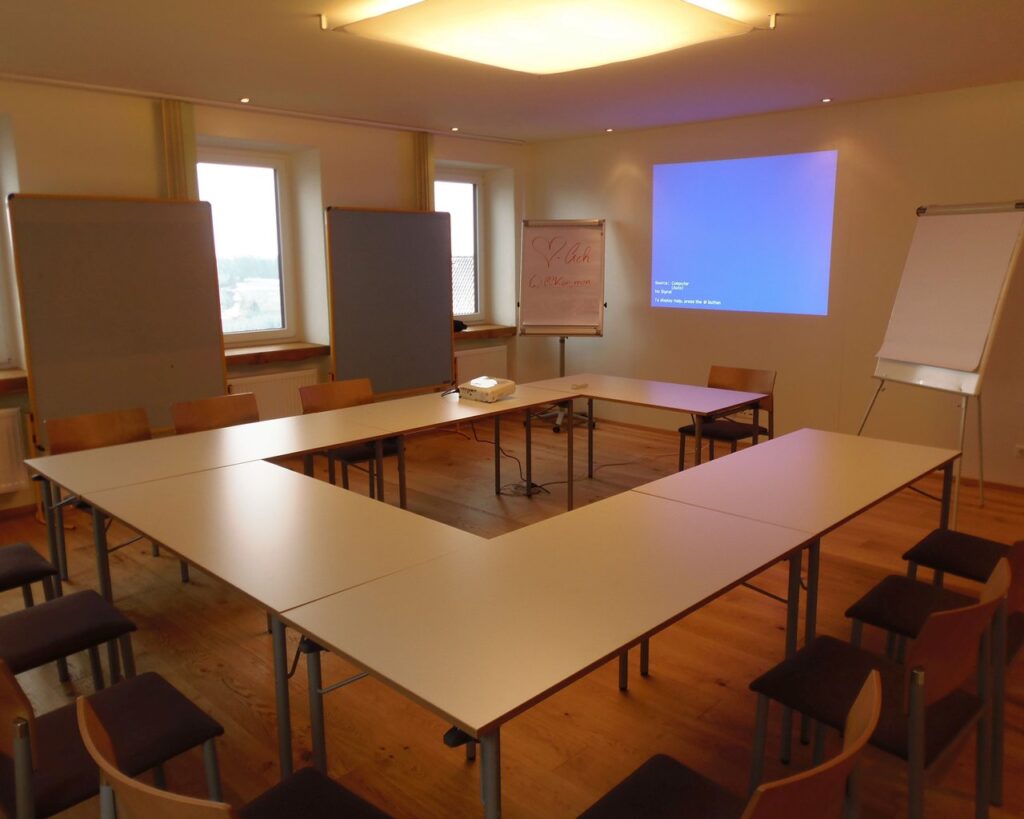 Konferenzraum mit großer Tischanordnung in U-Form, Stühlen, Flipcharts auf beiden Seiten und einem Projektorbildschirm an der Wand.