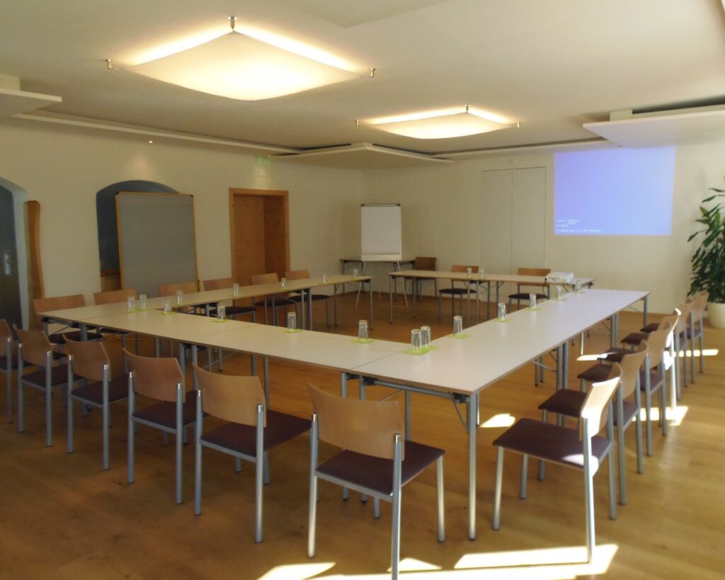 Konferenzraum mit rechteckiger Tischanordnung, umgeben von braunen Stühlen, und einem Projektorbildschirm an der vorderen Wand.