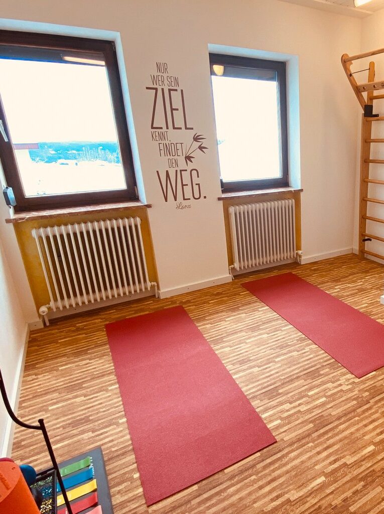 Heller Trainingsraum mit zwei Fenstern, darunter Heizkörper und roten Yogamatten auf dem Boden. An der Wand ein Plakat mit dem Spruch „Nur wer sein Ziel kennt, findet den Weg.“