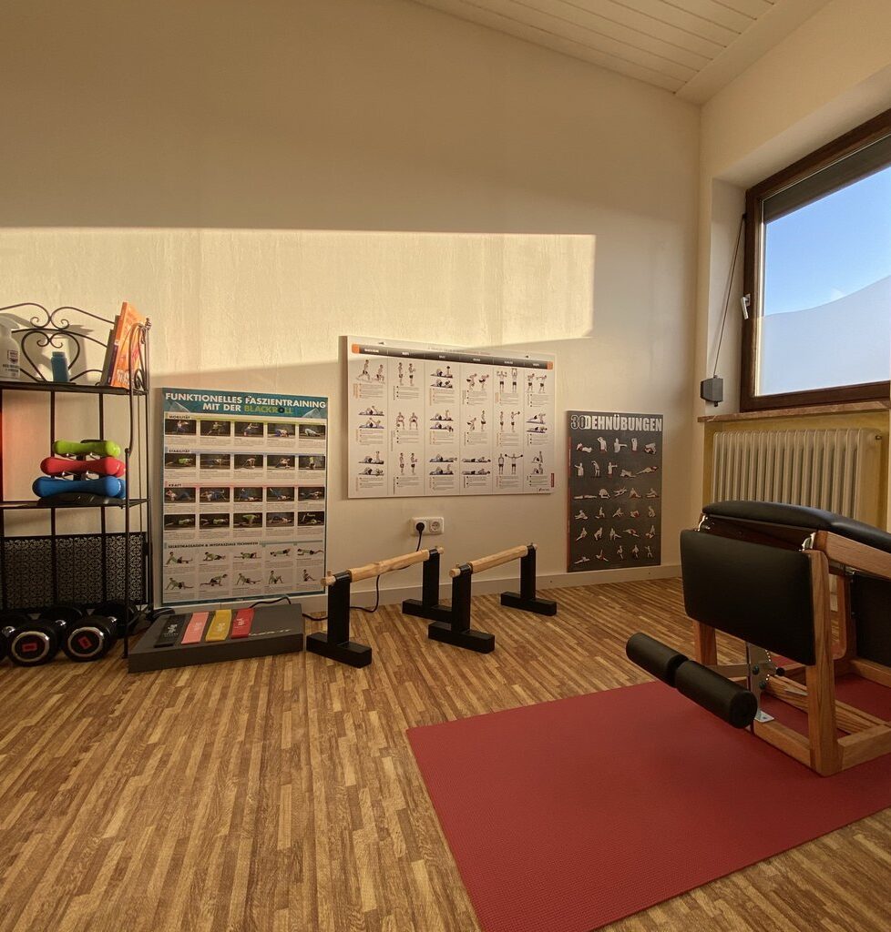 Innenansicht eines kleinen Fitnessraums mit einem Poster von Trainingsübungen an der Wand, Gewichten, einer Matte und einem Übungsgerät auf einem Holzboden.