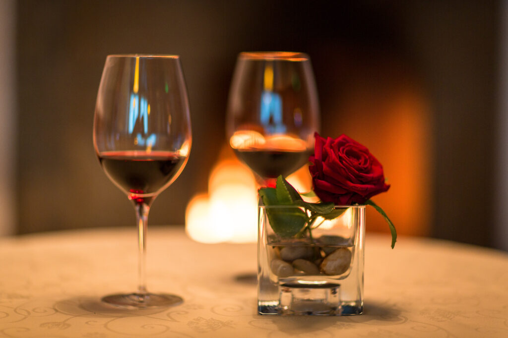 Zwei Weingläser und eine rote Rose in einem Glas auf einem Tisch vor unscharfem Hintergrund mit Kerzenlicht.