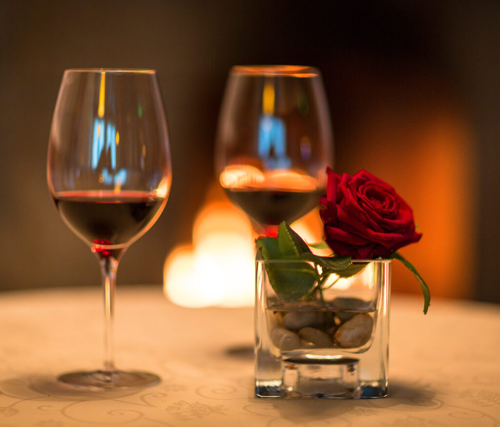 Zwei Weingläser gefüllt mit Rotwein, dazwischen eine rote Rose in einem Glas auf einem Tisch, im Hintergrund unscharf sichtbares Kerzenlicht.