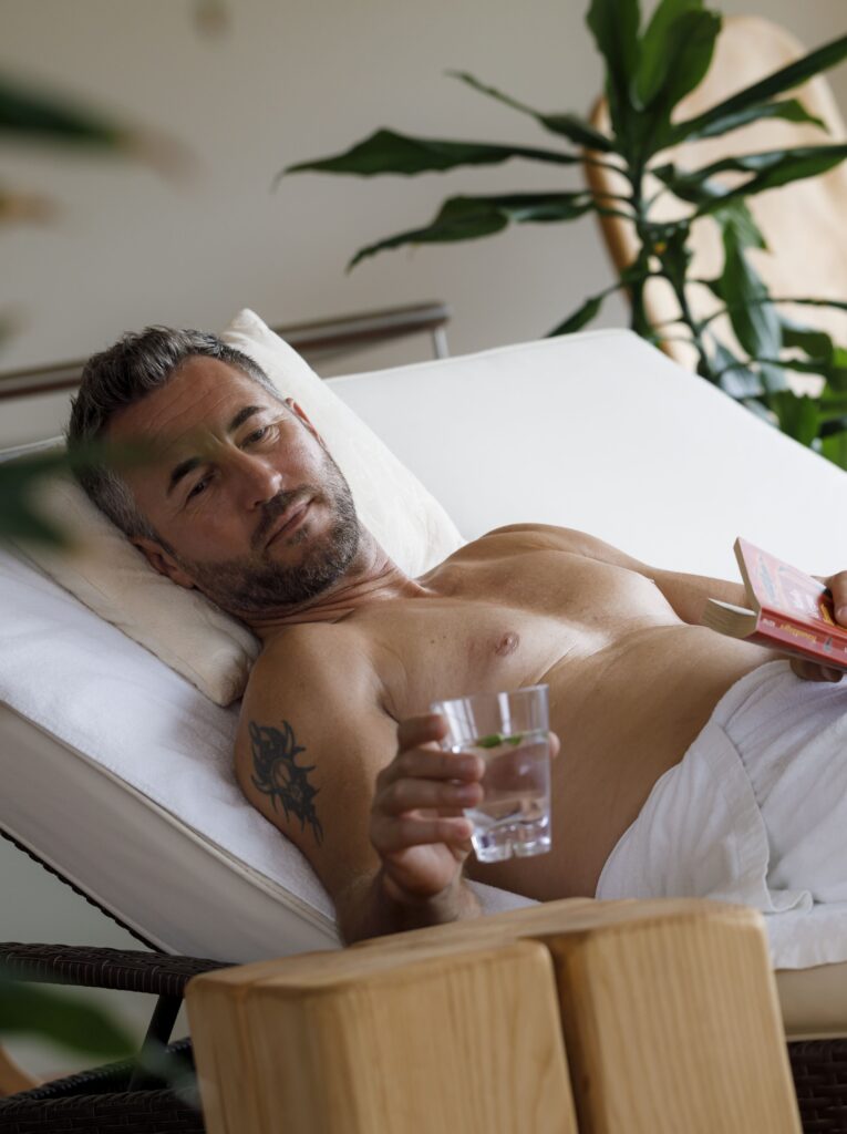 Ein entspannter Mann mittleren Alters mit grauem Haar und einem Tattoo am Arm liegt in einem weißen Handtuch gewickelt auf einer Liege, hält ein Glas Wasser und liest ein Buch neben einer großen Pflanze.