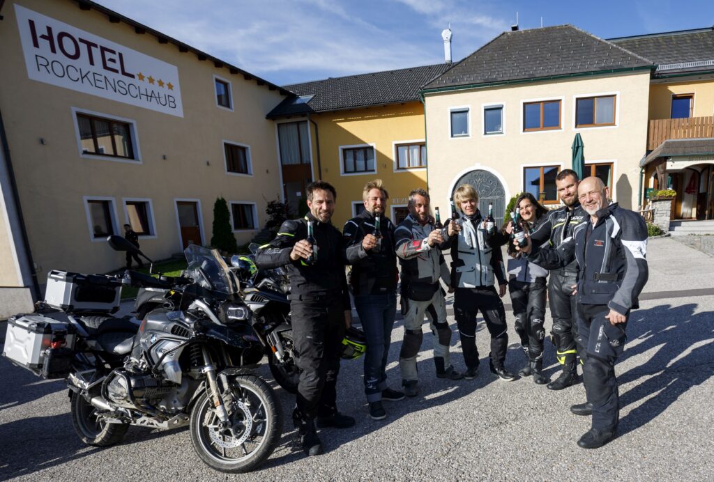 Gruppe von Motorradfahrern posiert vor einem Hotel namens "Hotel Rockenschaub" an einem sonnigen Tag.