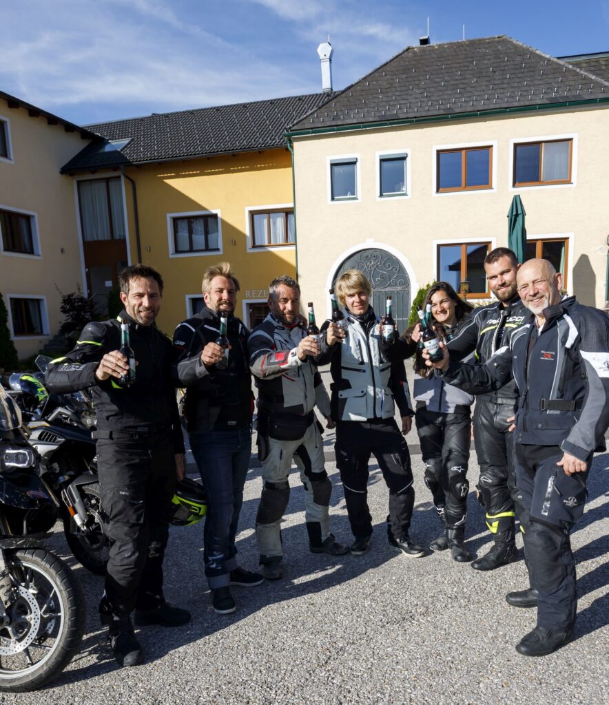 Gruppe von Motorradfahrern stößt mit Bierflaschen an, im Hof vor einem gelben Gebäude.