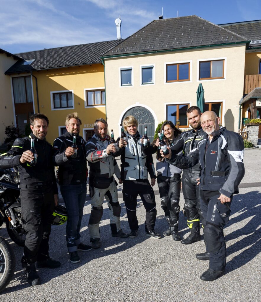 Gruppe von Motorradfahrern mit Helmen und Lederjacken, die vor einem gelben Gebäude stehen und zur Kamera lächeln, während sie ihre Gläser für einen Toast heben.