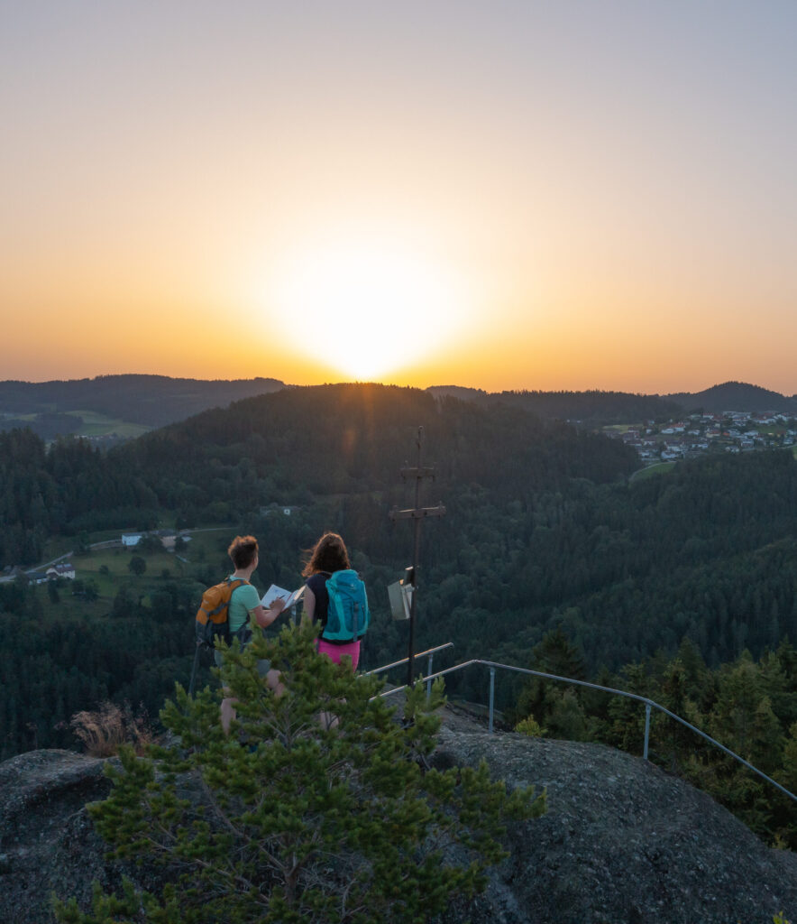 Zwei Personen sitzen auf einem Felsen und betrachten den Sonnenuntergang über einer bewaldeten Hügellandschaft.