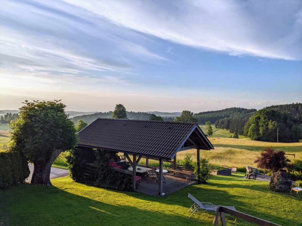 Ein idyllisches Landschaftsbild mit einem überdachten Picknickbereich im Vordergrund, umgeben von grünen Wiesen und Bäumen, mit einem sanft hügeligen Wald im Hintergrund unter einem klaren blauen Himmel.