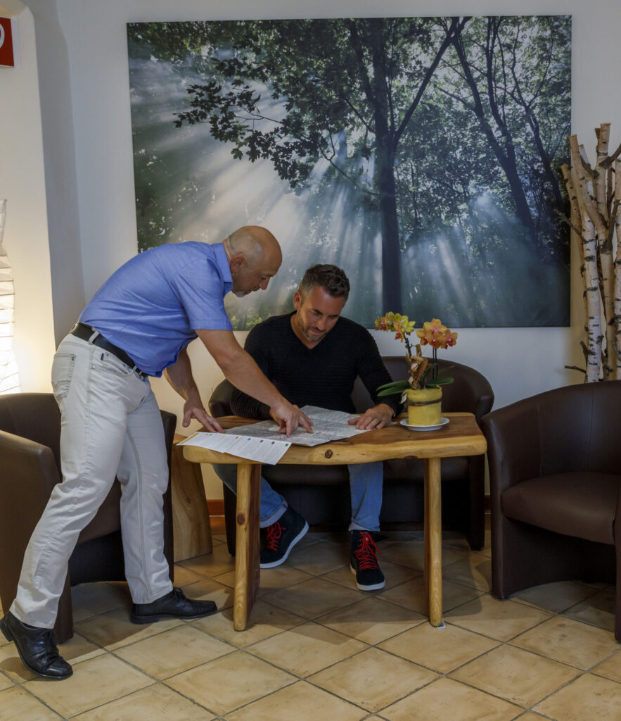 Zwei Männer betrachten einen Stadtplan an einem kleinen Holztisch in einem Café mit einer Waldlandschaft im Hintergrund.