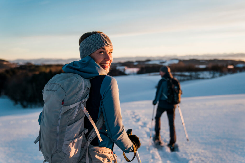Frau mit Rucksack und Mütze lächelt, während sie in einer winterlichen Landschaft mit Schneeschuhen wandert, im Hintergrund eine zweite Person.