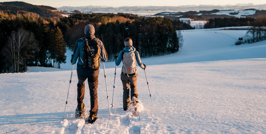 Zwei Personen wandern mit Schneeschuhen durch eine verschneite Landschaft bei Sonnenuntergang.
