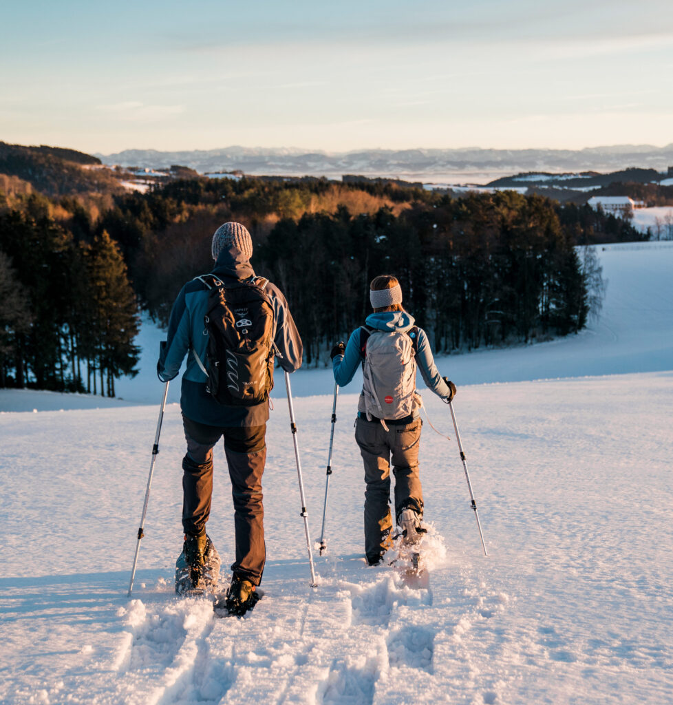 Zwei Personen wandern mit Schneeschuhen durch eine verschneite Landschaft bei Sonnenuntergang.