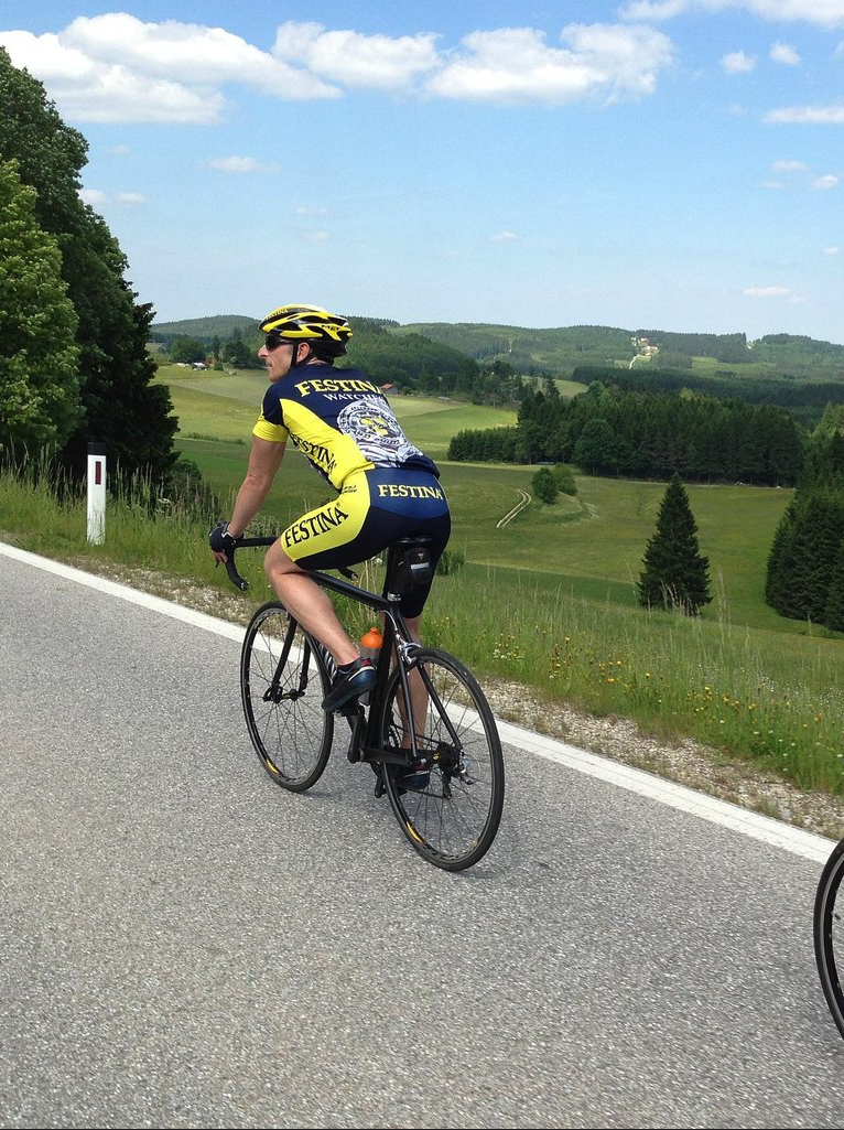 Ein Radfahrer in voller Montur fährt auf einer Landstraße. Im Hintergrund sind grüne Wiesen und bewaldete Hügel.