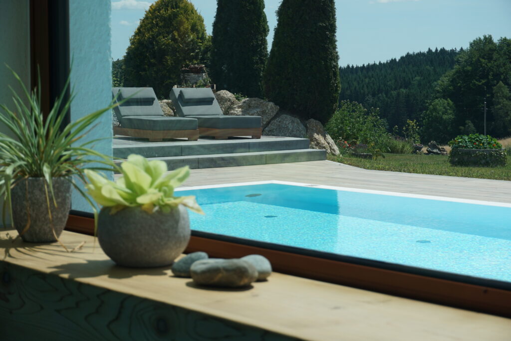 Blick durch ein Fenster auf einen Außenpool mit klarem blauem Wasser, umgeben von Liegestühlen und grünen Pflanzen, mit einem Wald im Hintergrund.