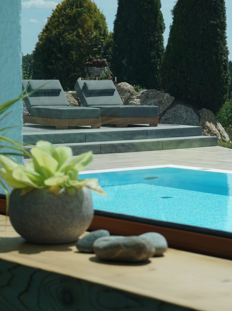 "Blick auf einen luxuriösen Außenpool mit Liegestühlen und einer grünen Gartenlandschaft im Hintergrund, teilweise verdeckt durch eine stilvolle Blumendekoration im Vordergrund."