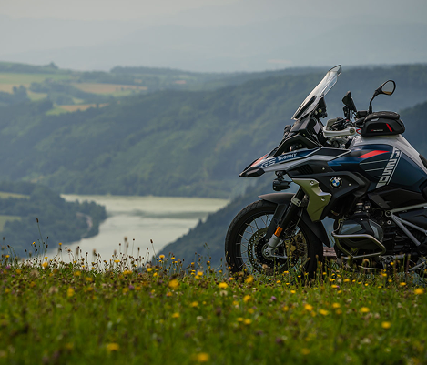 Ein BMW-Motorrad parkt auf einer grünen Wiese mit Blick auf ein Flusstal und bewaldete Hügel im Hintergrund.