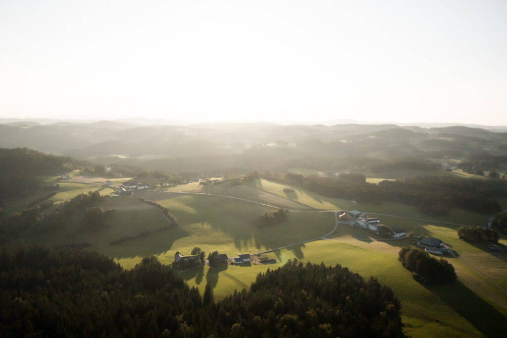 Landschaftsaufnahme aus der Vogelperspektive, die sanft hügelige Felder und kleine Siedlungen bei Sonnenuntergang zeigt.