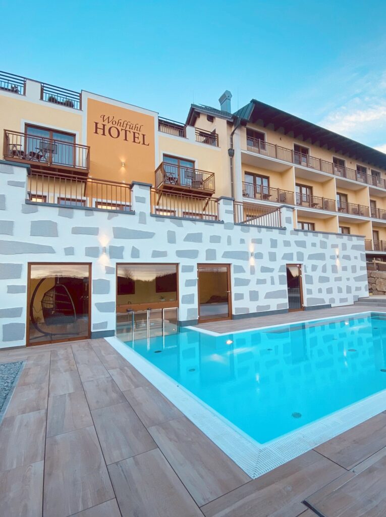 Außenansicht eines modernen Hotels mit beleuchtetem Swimmingpool im Vordergrund bei Dämmerung.