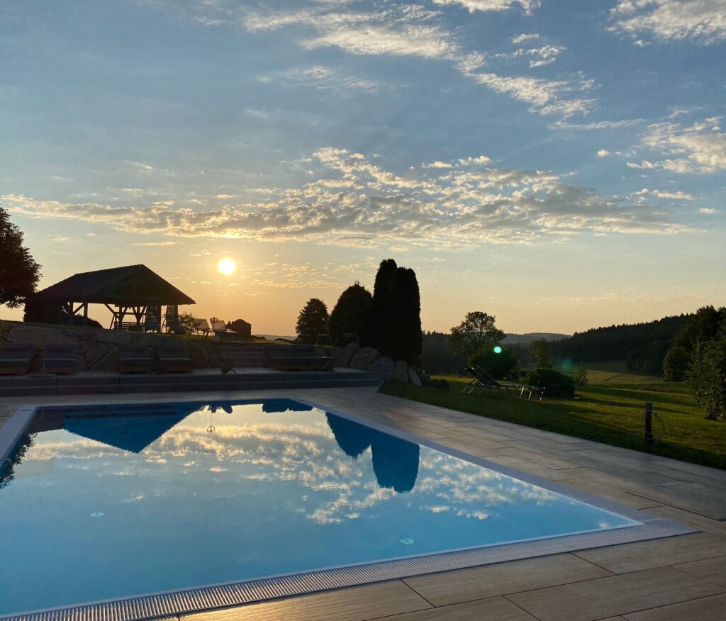 Sonnenuntergang spiegelt sich in einem Außenpool mit Blick auf grüne Hügel.