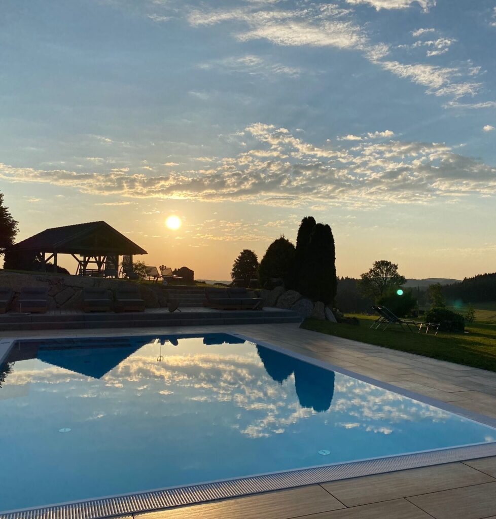 Sonnenuntergang über einem Swimmingpool mit Spiegelung des Himmels im Wasser, umgeben von einer Landschaft mit Bäumen.