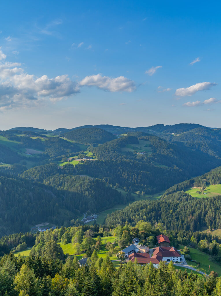 Blick auf eine idyllische Landschaft mit grünen Hügeln und Wäldern, im Vordergrund ein großes Gehöft, unter einem weiten, blauen Himmel mit einigen Wolken.