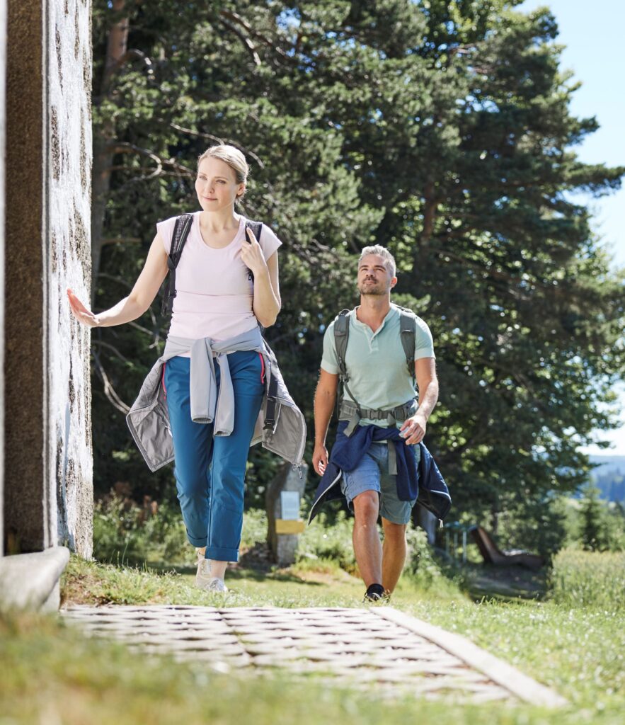 "Zwei Personen beim Wandern in einem sonnigen Waldgebiet, eine Frau und ein Mann in Freizeitkleidung."