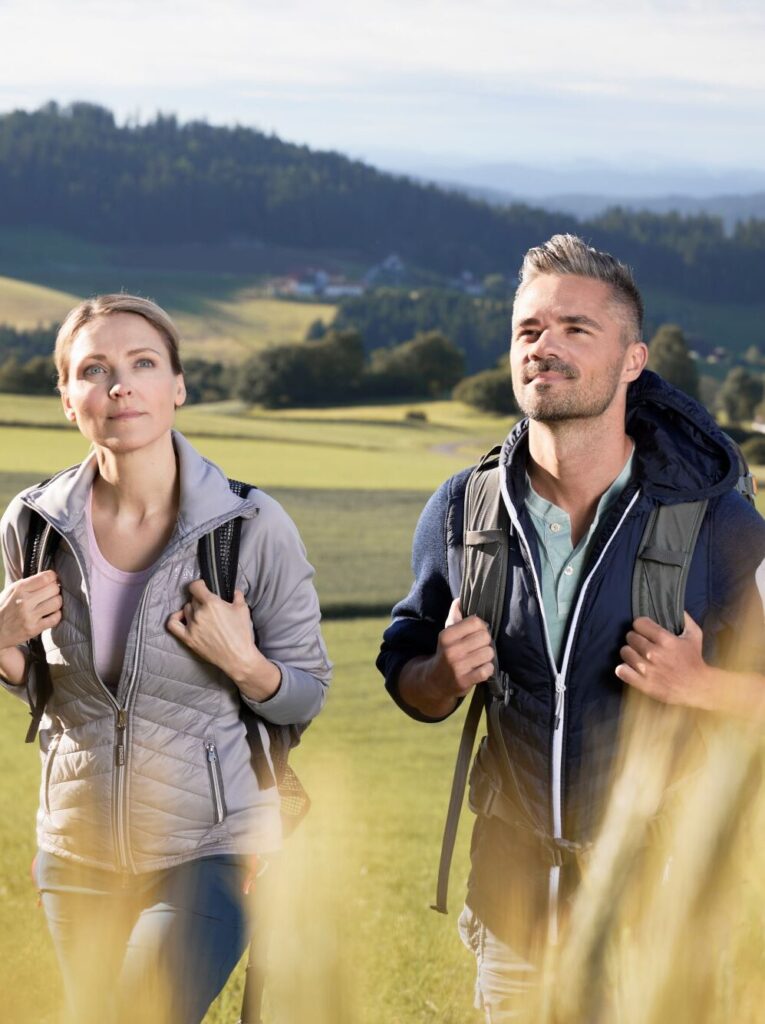 Ein Mann und eine Frau wandern bei sonnigem Wetter durch eine hügelige Landschaft.