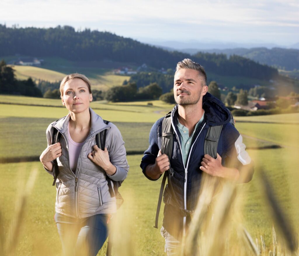 Ein Mann und eine Frau wandern bei sonnigem Wetter durch ein ländliches Gebiet mit grünen Hügeln im Hintergrund.