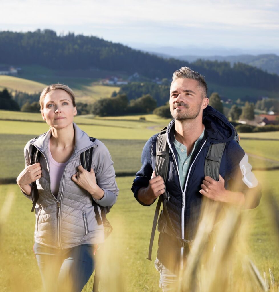 Ein Mann und eine Frau wandern bei sonnigem Wetter über eine Wiese im hügeligen Landschaft.
