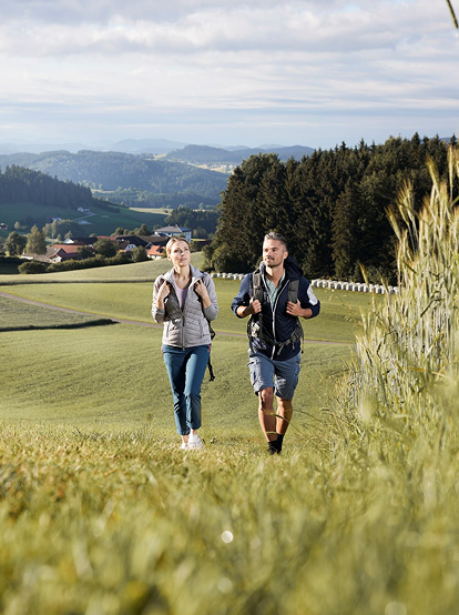 Ein Mann und eine Frau joggen auf einem Feldweg in einer ländlichen Gegend mit grünen Feldern und Hügeln im Hintergrund.