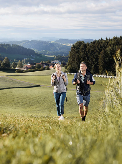 Ein junges Paar beim Wandern auf einem idyllischen Feldweg mit grünen Hügeln und einem bewaldeten Horizont im Hintergrund.
