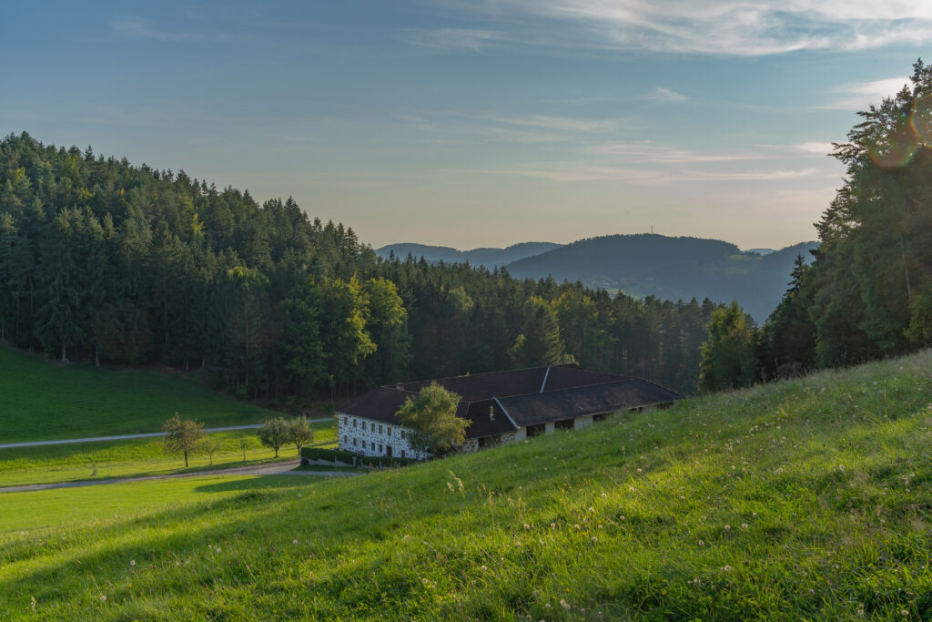 "Blick auf ein traditionelles Haus in einer ländlichen Gegend im Tal mit bewaldeten Hügeln im Hintergrund bei Sonnenuntergang."