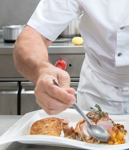Koch in weißer Uniform gibt mit einer Zange das letzte Stück Fleisch auf einen Teller mit fein angerichteten Speisen in einer professionellen Küche.