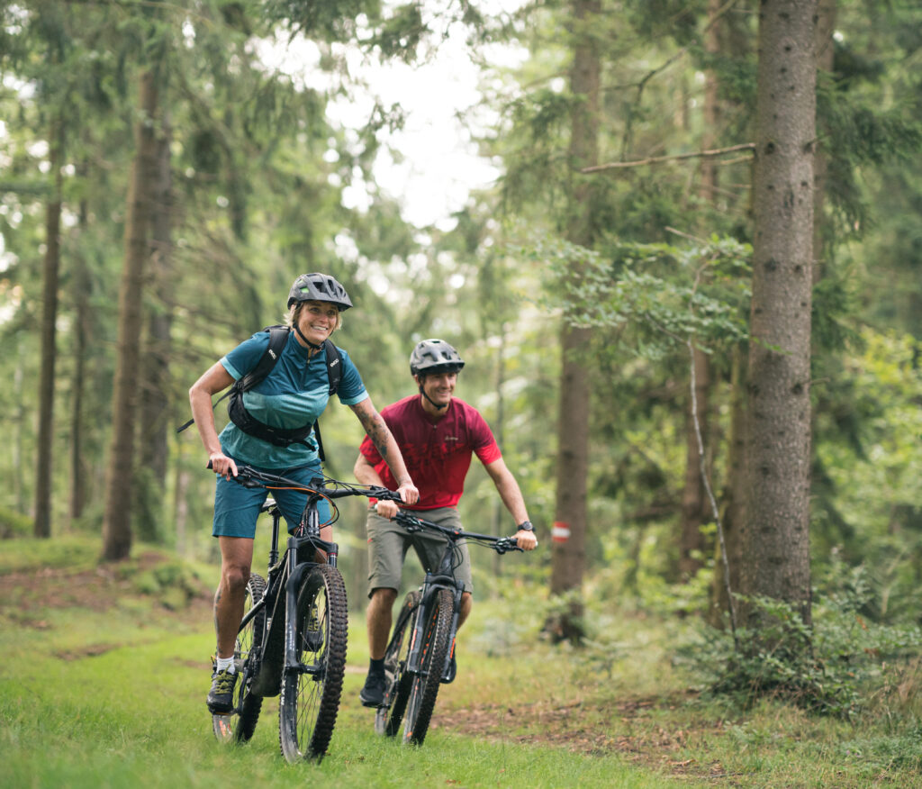 Zwei Personen fahren auf Mountainbikes durch einen Wald. Eine trägt ein blaues Shirt und die andere ein rotes Shirt. Beide tragen Helme.