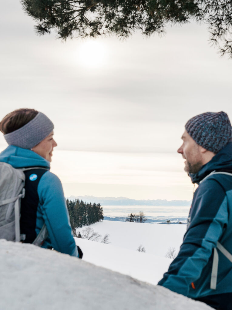 Zwei Personen in Winterkleidung sitzen auf einem schneebedeckten Hügel und blicken sich an, mit einer verschneiten Landschaft und Bergen im Hintergrund.