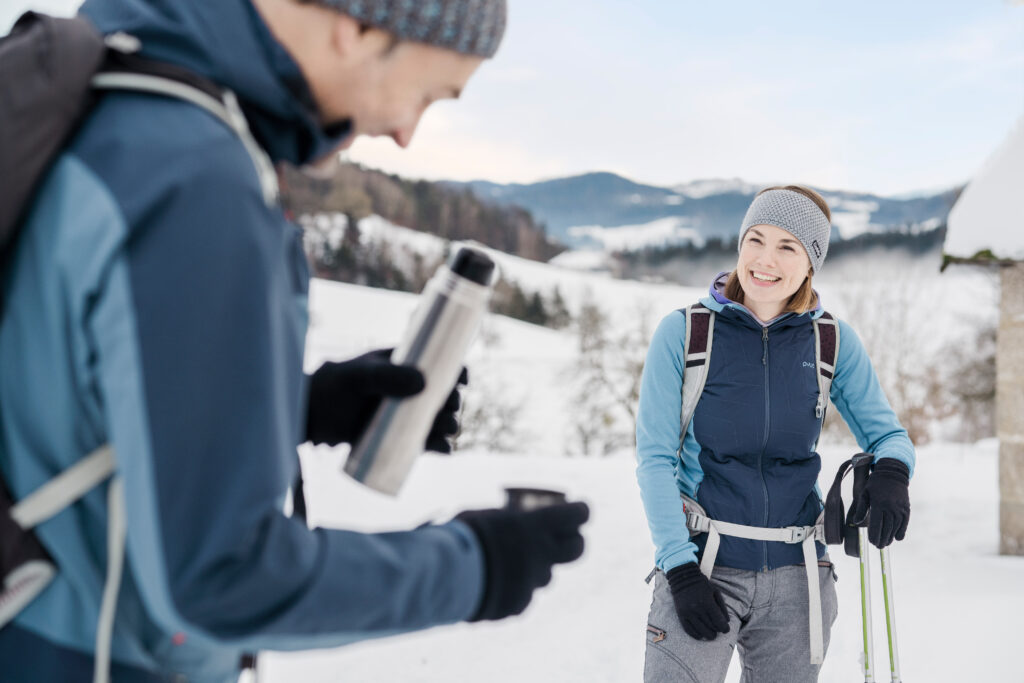 Ein Mann öffnet eine Thermoskanne und eine Frau mit Skistöcken lächelt ihn in einer schneebedeckten Landschaft an.