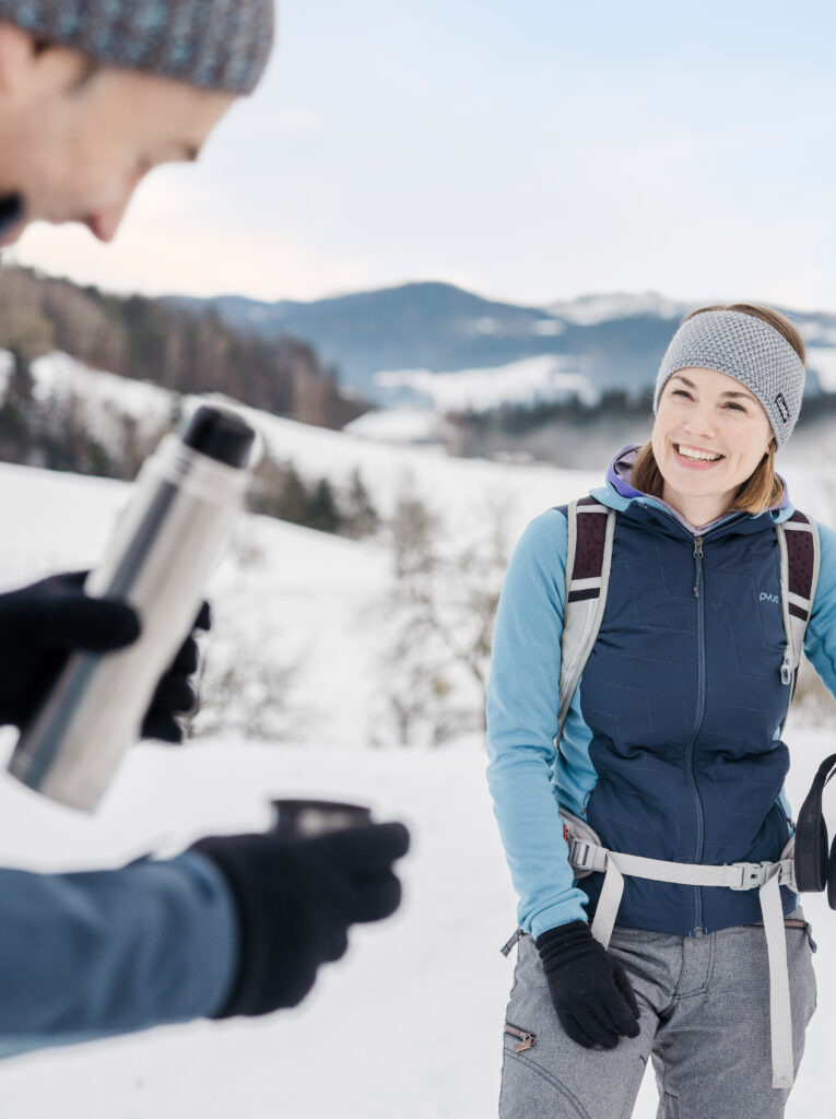 Frau erhält eine Thermosflasche von einer anderen Person im verschneiten Bergpanorama.