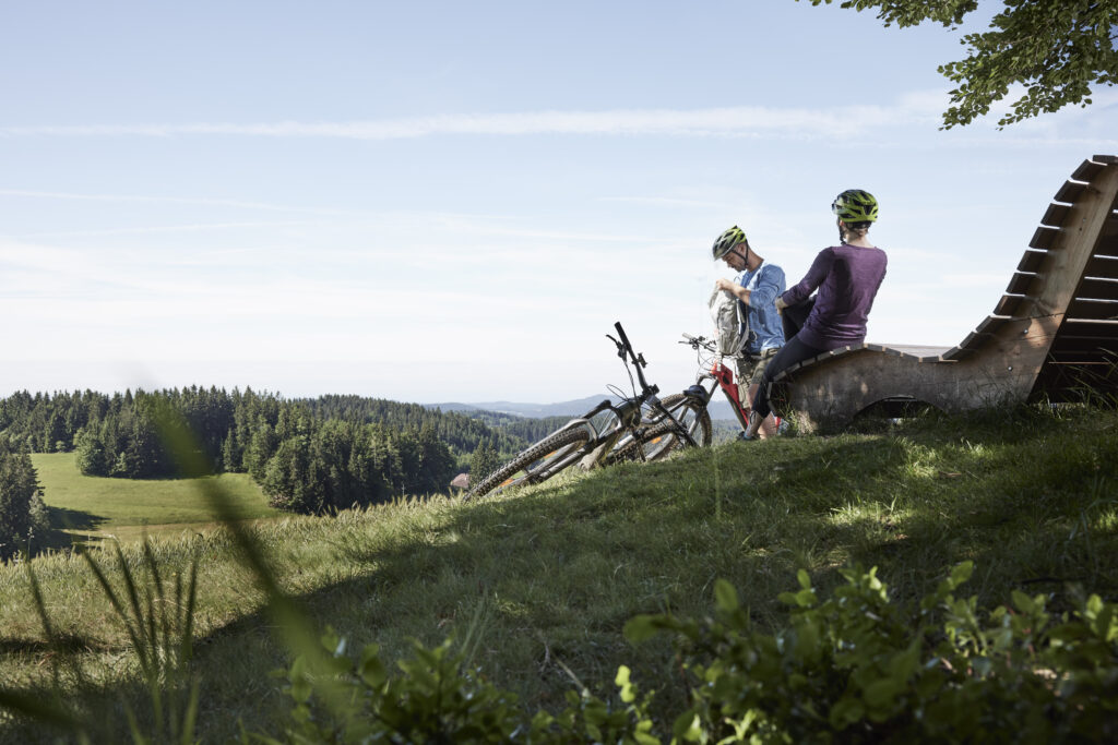 Zwei Personen in Fahrradhelmen sitzen auf einer Holzbank und betrachten die Aussicht in einem grünen Waldgebiet, ihre Mountainbikes neben sich abgestellt.