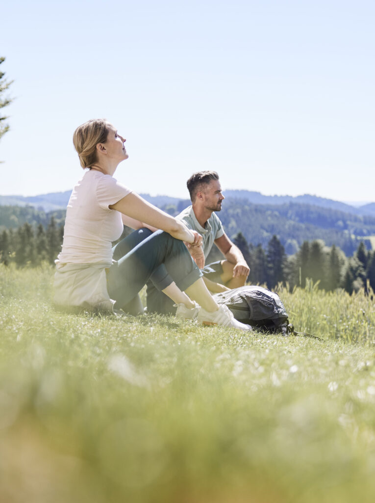 Ein Mann und eine Frau sitzen auf einer Wiese und blicken auf eine hügelige Landschaft. Sie tragen Freizeitkleidung und neben ihnen liegt ein Rucksack.