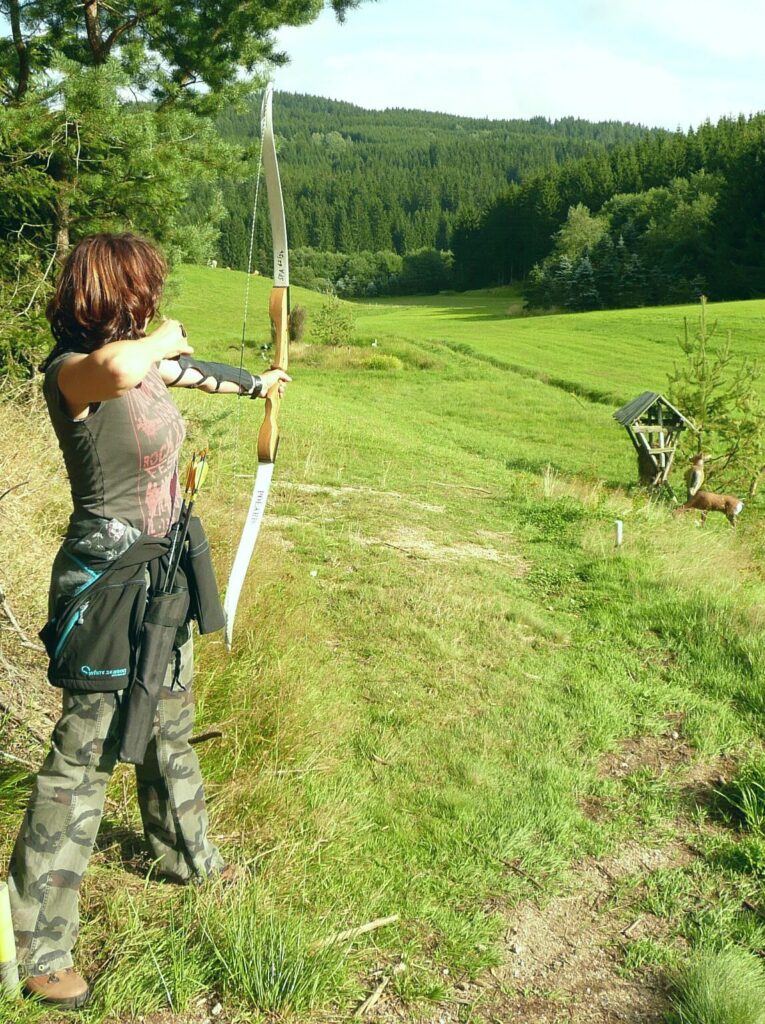 Eine Person beim Bogenschießen im Freien, mit einem großen Bogen, vor einer grünen Wald- und Wiesenlandschaft.