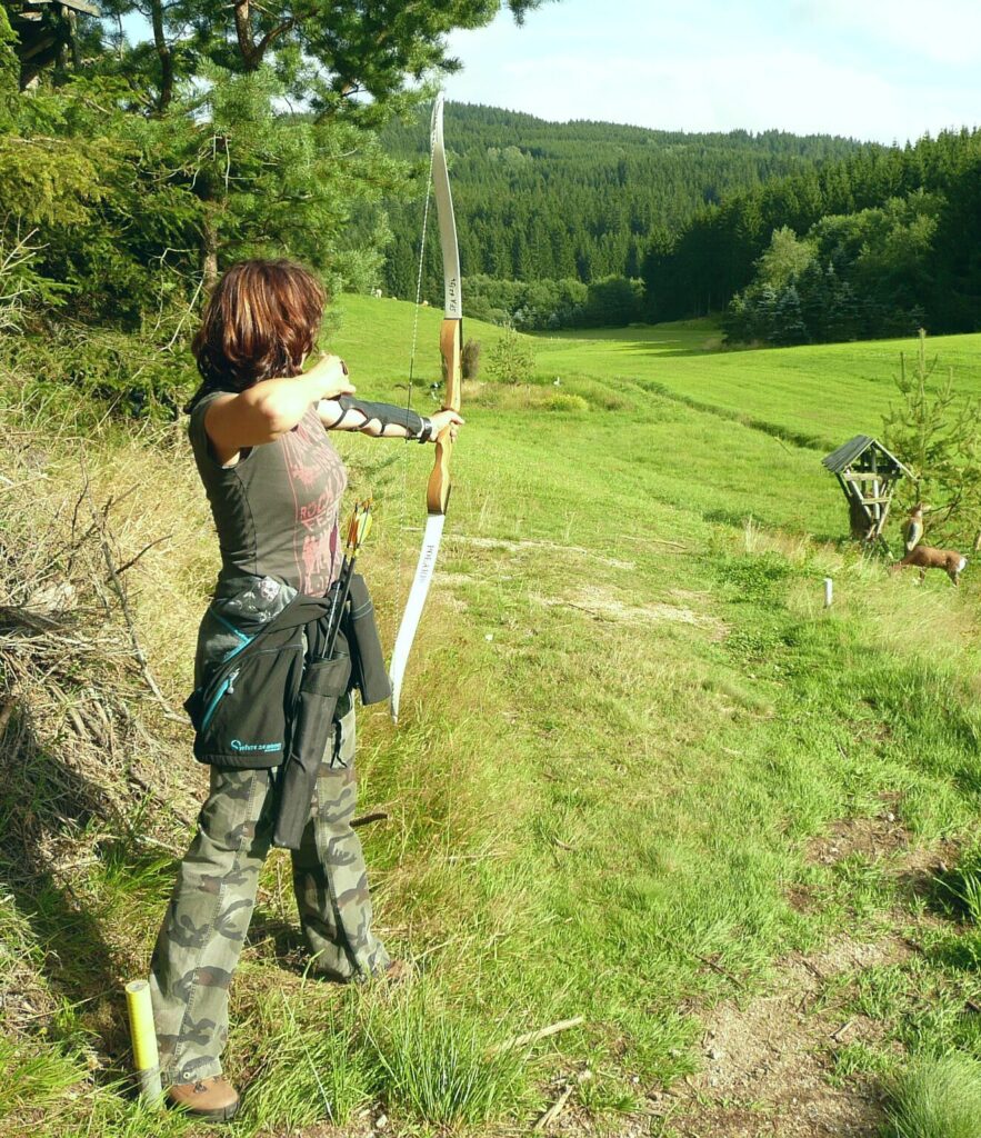 Frau beim Bogenschießen auf einer Wiese mit Waldlandschaft im Hintergrund.