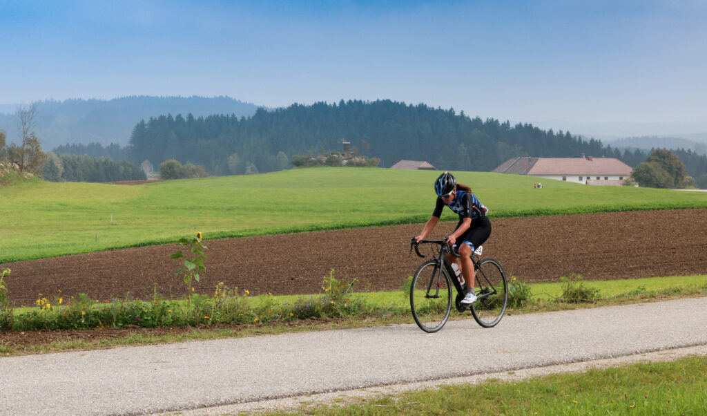 Ein Radfahrer fährt auf einem kleinen Weg neben einem frisch gepflügten Feld vor grünen Wiesen und einem bewaldeten Hügel im Hintergrund.