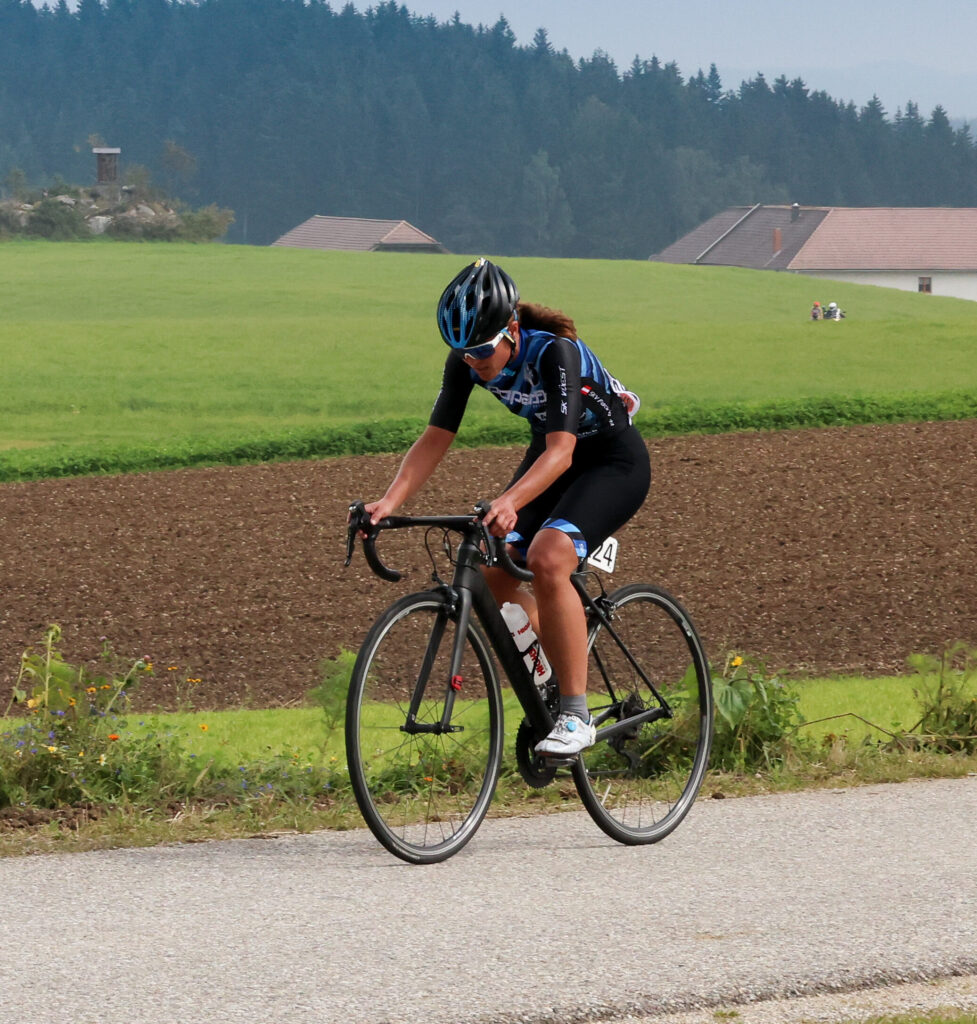 Radfahrer in Rennmontur fährt auf einem Landweg mit Feldern im Hintergrund.