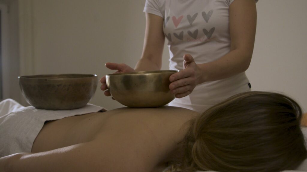 Eine Person erhält eine Klangschalentherapie, wobei zwei Klangschalen auf ihrem Rücken stehen, während eine Therapeutin sie hält.