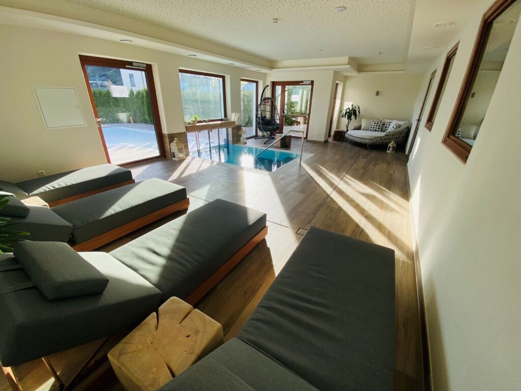 Helles Wohnzimmer mit großen Fenstern, modernen grauen Sofas und hölzernem Fußboden, mit Blick auf einen Poolbereich.