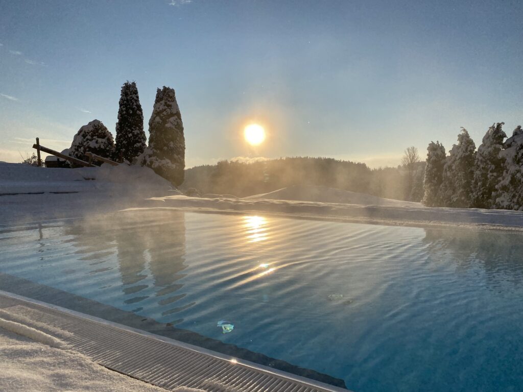 Schneebedeckte Landschaft mit einem beheizten Außenpool unter einem klaren, sonnigen Himmel bei Sonnenuntergang.