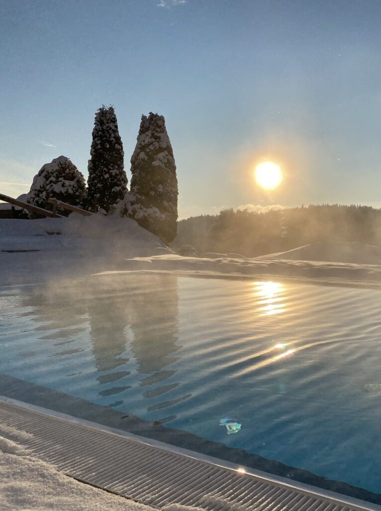 Ein sonniger Wintertag an einem Außenpool mit dampfendem Wasser, umgeben von schneebedeckten Tannen und einem klaren blauen Himmel.