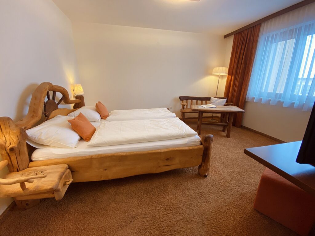 "Hotelzimmer mit rustikalem Holzbett, Nachttisch, Schreibtisch und Fernseher. Warmes, einladendes Ambiente mit Teppichboden und weicher Beleuchtung."